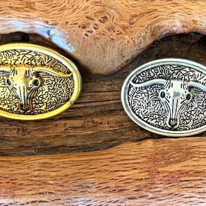 May include: Two oval belt buckles with a bull skull design. One is gold-toned, the other silver-toned. Both have intricate floral patterns around the skull. The buckles are displayed on a textured wooden surface.
