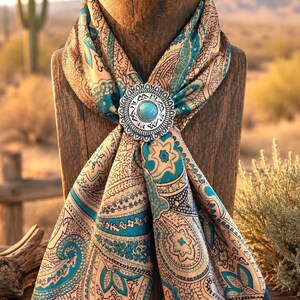 May include: A silk scarf featuring a teal, beige, and black paisley pattern, secured with a circular silver-toned metal slide. The slide has a scalloped edge, engraved geometric details, and a central round turquoise-colored stone inlay.