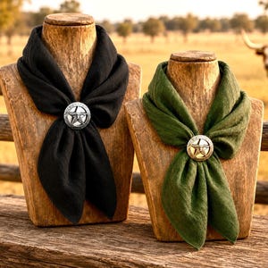 May include: Two neckerchiefs, one black and one olive green, each secured with a circular silver-toned slide featuring a star emblem. The fabric has a textured appearance, and the scarves are draped over wooden display forms.