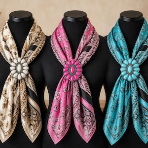 May include: Three scarves with floral patterns in cream, pink, and teal, each secured with a large oval concho-style slide featuring a central stone surrounded by smaller stones in matching colours, displayed on black mannequin torsos.
