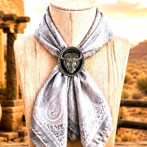 May include: A light gray silk scarf featuring a dark gray paisley pattern, secured with a circular metal slide. The slide displays a detailed, raised relief of a bison head in a dark metallic finish, centered on the fabric folds.