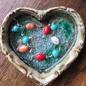 May include: Two bracelets with turquoise and coral stones on a heart-shaped dish with a mosaic of green and white glass.