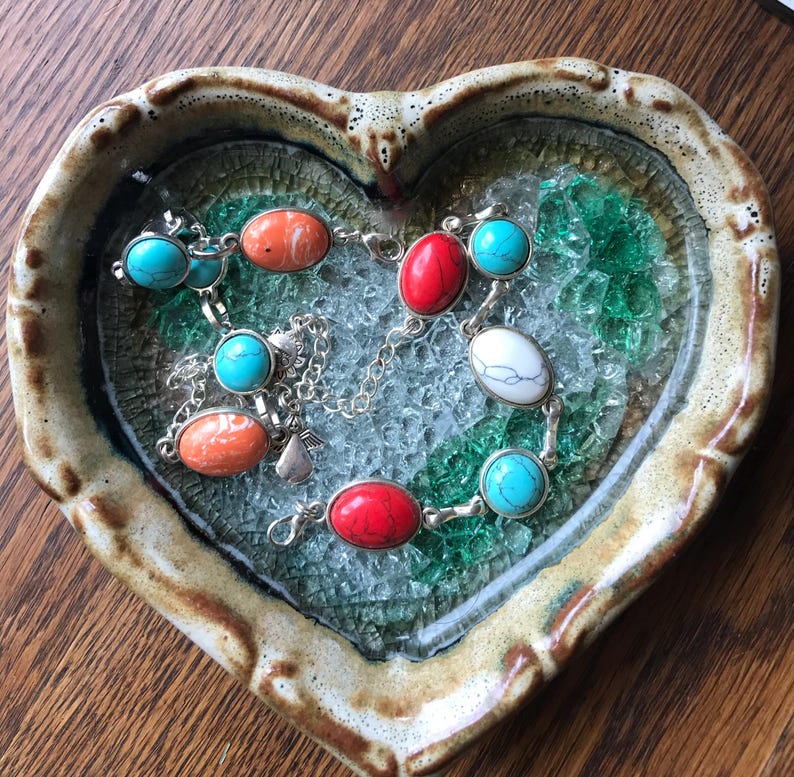 May include: Two bracelets with turquoise and coral stones on a heart-shaped dish with a mosaic of green and white glass.