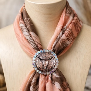 May include: A peach and brown patterned scarf secured with a circular copper-toned scarf slide. The slide features an embossed bull skull design in the center, surrounded by a border of small, iridescent, multi-colored faceted crystals.