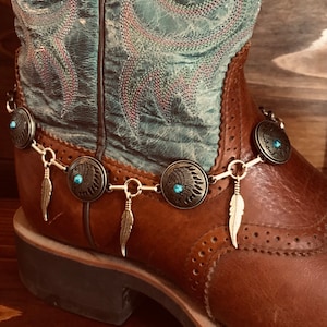 May include: A pair of brown leather cowboy boots with a turquoise and gold beaded ankle bracelet. The bracelet features small feather charms and Native American headdress charms.
