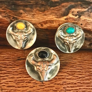 May include: Three silver bull skull pendants with different colored stones. The stones are yellow, turquoise, and black.