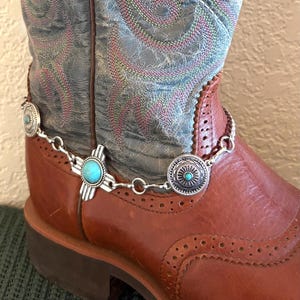 Puede incluir: Bota de vaquero de cuero marrón con costuras decorativas y una cadena de bota plateada con detalles turquesa y plateados. La bota tiene una parte superior bordada en azul y rosa desgastado.