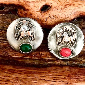 Op de afbeelding: Twee zilverkleurige metalen knopen met een hoefijzer- en paardendesign. De ene knop heeft een groene steen en de andere een rode steen.