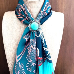 May include: A teal scarf with a floral pattern and a turquoise brooch. The scarf is draped over a mannequin, highlighting the design and the brooch's silver filigree. The scarf's pattern includes pink and white floral elements.