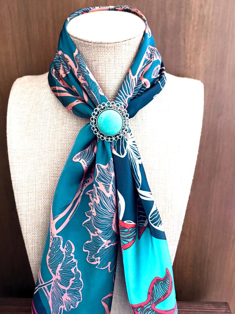 May include: A teal scarf with a floral pattern and a turquoise brooch. The scarf is draped over a mannequin, highlighting the design and the brooch's silver filigree. The scarf's pattern includes pink and white floral elements.