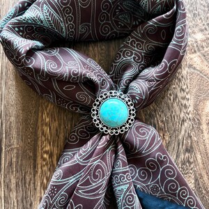 May include: A brown scarf with a teal paisley pattern and a turquoise and silver bolo tie. The scarf is folded and draped, showcasing the intricate design. The bolo tie is round with a bright blue stone.