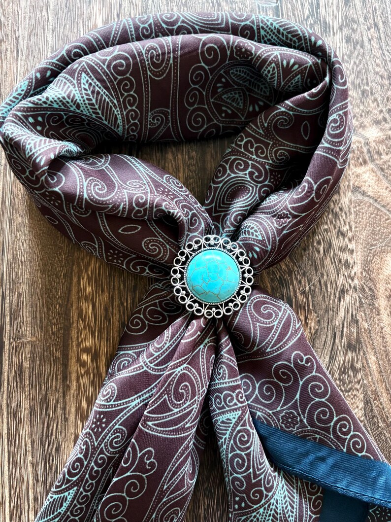May include: A brown scarf with a teal paisley pattern and a turquoise and silver bolo tie. The scarf is folded and draped, showcasing the intricate design. The bolo tie is round with a bright blue stone.