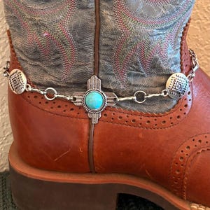 Op de afbeelding: Close-up van een bruine leren cowboylaars met een decoratieve zilveren ketting. De ketting heeft turquoise en zilveren accenten, waaronder een turkooizen steen in het midden. De laars heeft een decoratief patroon op de schacht.