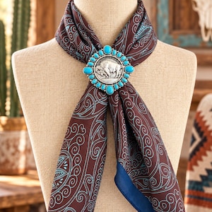 May include: A brown silk scarf featuring a light blue swirling pattern, secured with a silver-toned metal slide. The slide is oval-shaped, decorated with a central embossed buffalo figure surrounded by a border of small, round turquoise stones.