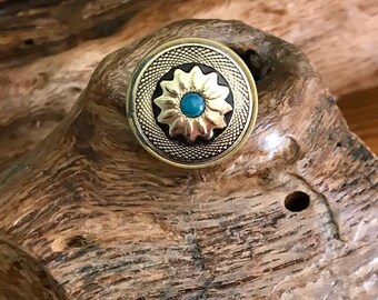 May include: A round, antique-style button with a gold-colored floral design and a small turquoise center. The button has a textured, silver-colored outer ring and is set against a natural wood background.