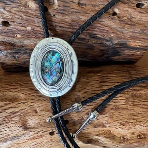 May include: Oval bolo tie with a silver-toned frame and a central abalone shell inlay. The shell displays iridescent blues and greens. The black leather cord has decorative silver tips and a sliding clasp. The bolo tie rests on a wooden surface.