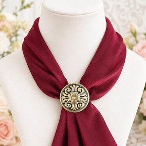May include: A deep burgundy scarf secured with a circular gold-toned metal scarf ring. The metal ring features an embossed floral pattern with intricate swirling details and a central flower motif, holding the fabric in a gathered knot style.