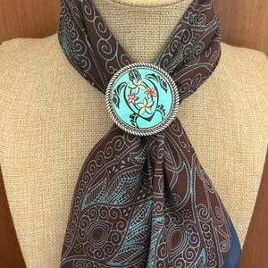 Puede incluir: Bufanda marrón con un diseño floral turquesa y blanco, sujeta por un pasador ovalado plateado. El pasador presenta un fondo turquesa con un diseño de tortuga y flores.