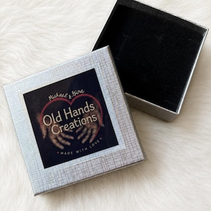 May include: A square silver gift box with a textured finish and an open lid. The top of the box features a label with the text Michael & Gina Old Hands Creations Made With Love, accompanied by an illustration of two hands forming a heart shape.