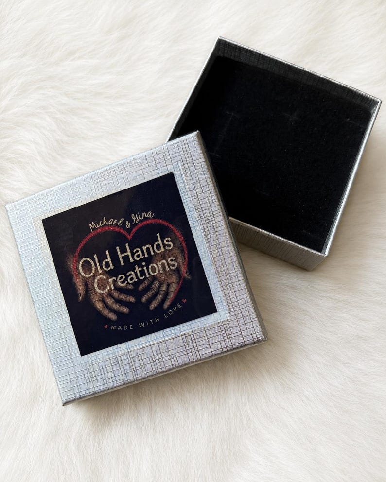 May include: A square silver gift box with a textured finish and an open lid. The top of the box features a label with the text Michael & Gina Old Hands Creations Made With Love, accompanied by an illustration of two hands forming a heart shape.