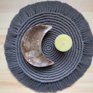 May include: A gray macrame coaster with a fringe border. A wooden crescent moon bowl and a yellow candle are placed on top of the coaster.