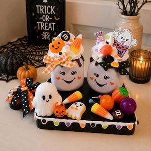 May include: Two white mugs with ghost faces, topped with faux whipped cream, candy corn, and pumpkin decorations. The mugs sit on a black tray decorated with ghost figures, colorful candy pieces, and a bow with orange and black polka dots.