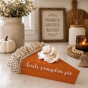 May include: A triangular wooden pumpkin pie slice decoration painted orange with the text fresh pumpkin pie in white script. It features a brown and white gingham fabric ruffle along the back edge and a white dollop of whipped cream on top.