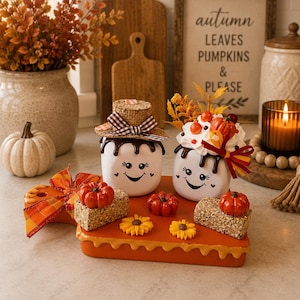 May include: Two white marshmallow-shaped figurines with smiling faces sit on an orange rectangular base. One wears a burlap hat, the other has faux whipped cream and berries. The base is decorated with two small hay bales, pumpkins, and flowers.