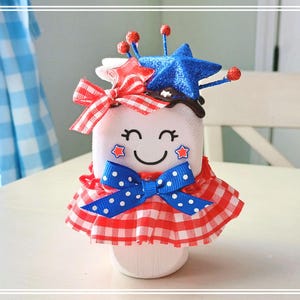 May include: A white ceramic figurine with a red, white, and blue gingham dress, a blue star, and red and white stars on its face. The figurine is smiling and has a red, white, and blue bow.