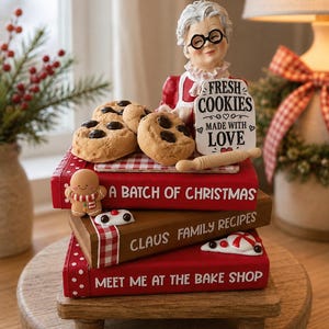 May include: A decorative figurine of an elderly woman with glasses holding a sign that reads FRESH COOKIES MADE WITH LOVE, placed atop a stack of three books titled A BATCH OF CHRISTMAS, CLAUS FAMILY RECIPES, and MEET ME AT THE BAKE SHOP, with cookies.