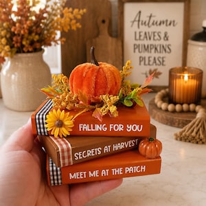 May include: A stack of three decorative wooden books in shades of orange and brown. The spines feature the text "FALLING FOR YOU," "SECRETS AT HARVEST," and "MEET ME AT THE PATCH." The stack is topped with a fuzzy orange pumpkin and autumn foliage.