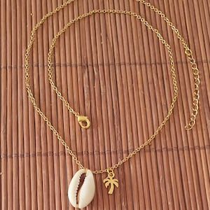 Könnte beinhalten: Eine goldene Kettenkette mit einem weißen Muschel-Anhänger und einem goldenen Palmen-Anhänger.