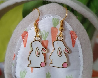 Puede incluir: Un par de pendientes de aro dorados con dijes de conejo blanco y detalles en rosa. Los dijes de conejo tienen un diseño simple y de dibujos animados.