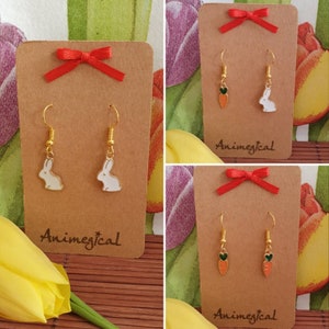 Könnte beinhalten: Zwei Paar Ohrringe auf braunen Karten mit roten Schleifen. Ein Set zeigt weiße Hasenanhänger und das andere Set zeigt orangefarbene Karottenanhänger mit grünen Spitzen. Die Karten tragen den Schriftzug "Animegical".