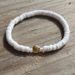 Könnte beinhalten: Ein elastisches Armband bestehend aus zahlreichen kleinen, flachen, kreisförmigen weißen Tonperlen. Das Armband verfügt über einen einzelnen goldfarbenen Metallanhänger in Form einer Muschel, flankiert von zwei kleinen, runden, weißen Perlen.