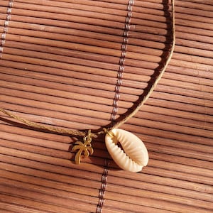 Könnte beinhalten: Eine goldene Halskette mit einem kleinen Palmenbaum-Anhänger und einem größeren Muschel-Anhänger. Die Halskette liegt auf einem braunen gewebten Hintergrund.