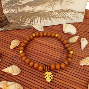Könnte beinhalten: Ein braunes Holz-Armband mit goldenen Perlen und einem goldenen Blattanhänger. Das Armband liegt auf einer braunen Bambusmatte mit Muscheln.