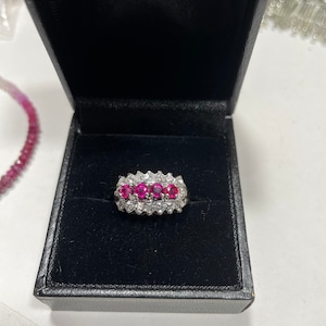 Pode incluir: Um anel de prata com uma fileira de pedras preciosas cor-de-rosa cercadas por pedras preciosas transparentes menores.