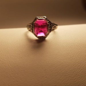 Puede incluir: Un anillo vintage con una piedra preciosa rosa engastada en una banda de oro. El anillo tiene detalles intrincados alrededor de la piedra.