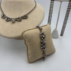 Può includere: Un set di gioielli con collana, bracciale e orecchini pendenti argentati. La collana e il bracciale presentano motivi floreali con pietre chiare e scintillanti. Gli orecchini sono lunghi, con più fili delle stesse pietre. I gioielli sono esposti su un busto e un cuscino color crema.