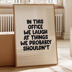 May include: A framed print with the text "IN THIS OFFICE WE LAUGH AT THINGS WE PROBABLY SHOULDN'T" in bold black letters. The print has a light beige background and a thin, brown frame. The image is set against a neutral-toned room.