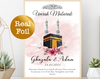 Op de afbeelding: Een roze en witte aquarel achtergrond met een zwarte en gouden illustratie van de Kaaba. De tekst "Umrah Mubarak" is geschreven in gouden kalligrafie boven de illustratie. Onder de illustratie staat de tekst "Ghazala & Adam" in zwarte kalligrafie, gevolgd door de datum "24.01.2023". De tekst "Moge je Umrah worden geaccepteerd. Moge al je Duas worden verhoord. Accepteer je daden van aanbidding. Versterk je geloof. Amin" is in zwarte tekst onder de datum geschreven.