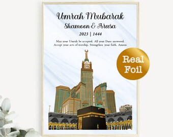 Op de afbeelding: Een digitale illustratie van de Kaaba in Mekka, Saoedi-Arabië. De afbeelding is omlijst met een houten rand en heeft een sticker van echt goudfolie met de tekst "Real Foil". De tekst "Umrah Mubarak Shamoon & Areesa 2023 | 1444" is onder de afbeelding gedrukt. De tekst "Moge je Umrah worden geaccepteerd. Moge al je Duas worden verhoord. Accepteer je daden van aanbidding. Versterk je geloof. Amin" is onder de datum gedrukt.