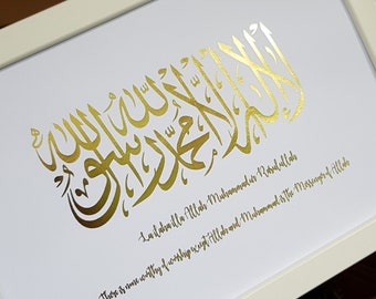 May include: A framed print with gold calligraphy in Arabic script. The text reads "La ilaha illa Allah Muhammadur Rasulullah" and "There is none worthy of worship except Allah and Muhammad is the Messenger of Allah".