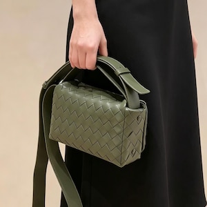 Può includere: Una borsa a mano rettangolare color verde oliva caratterizzata da un motivo in pelle intrecciata. La borsa include una spessa tracolla in tinta verde oliva e un manico superiore, con una forma strutturata e dettagli di cuciture laterali.
