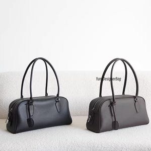 May include: Two leather handbags, one black and one dark gray, with matching handles and zipper closures. Each bag has a rectangular shape and a small tag attached to the zipper. The bags are displayed on a white, textured surface.