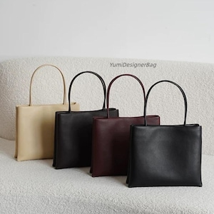 May include: Four rectangular handbags with thin top handles, arranged in a row. The bags are colored beige, black, dark brown, and black. Each bag features a smooth finish and a structured shape. The text YumiDesignerBag is visible above them.