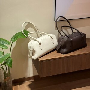 May include: Two leather barrel-shaped handbags. One is off-white, the other is dark brown. Both have top handles and zippers. They are displayed on a wooden surface against a neutral wall.