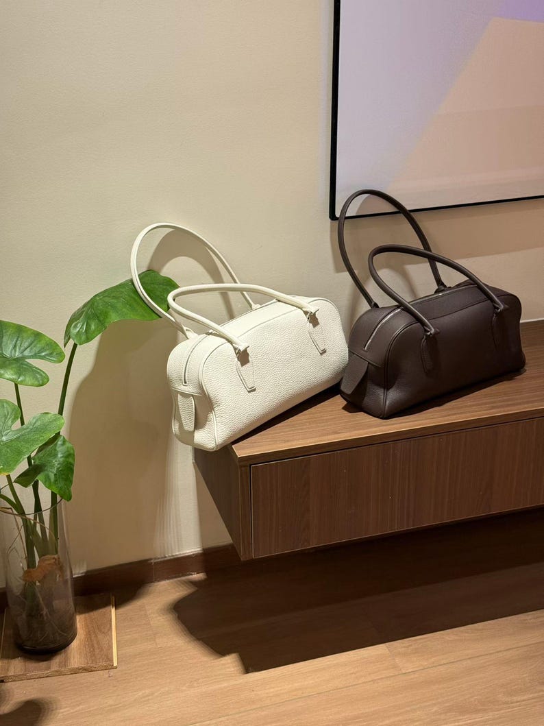 May include: Two leather barrel-shaped handbags. One is off-white, the other is dark brown. Both have top handles and zippers. They are displayed on a wooden surface against a neutral wall.
