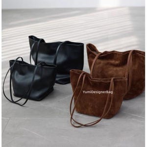 Peut inclure: Quatre sacs fourre-tout de différentes tailles et couleurs. Deux sacs en cuir noir et deux sacs en daim marron avec de longues anses sont présentés. Les sacs sont sur une surface gris clair. Le texte "YumiDesignerBag" est visible.